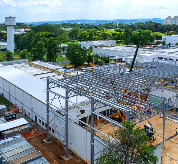 Obra vista do alto, com estrutura metálica de cobertura em montagem, guindaste e área verde ao redor